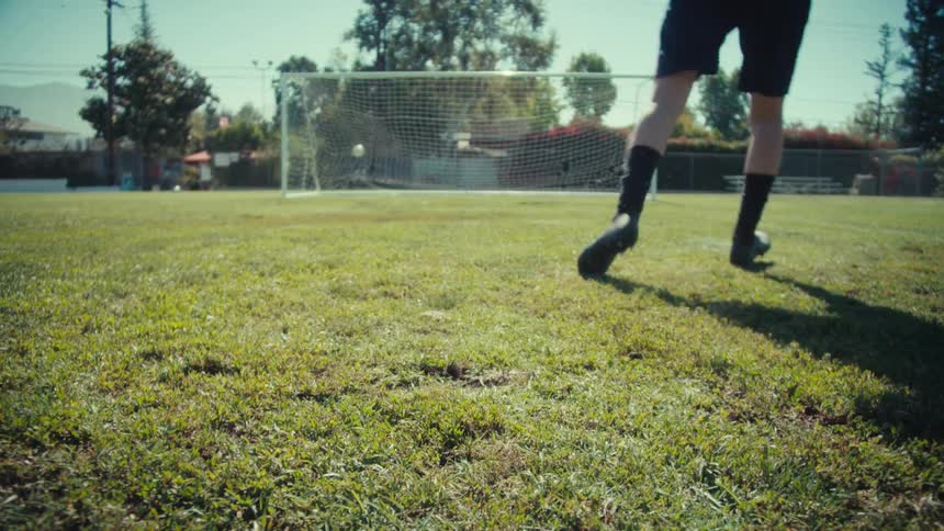 From a low angle, a soccer player runs up and kicks a ball, scoring a goal on a sunny green field