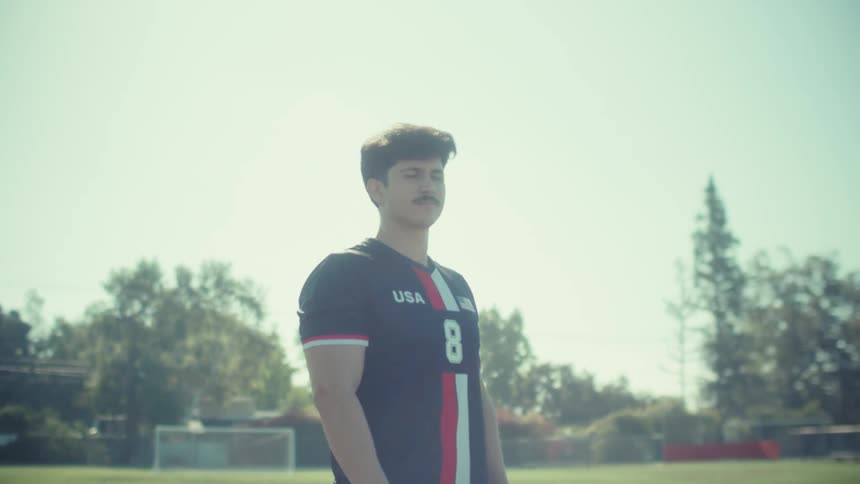 A determined young male soccer player in a USA jersey stands proudly on a sunny field in a slow-motion push-in shot