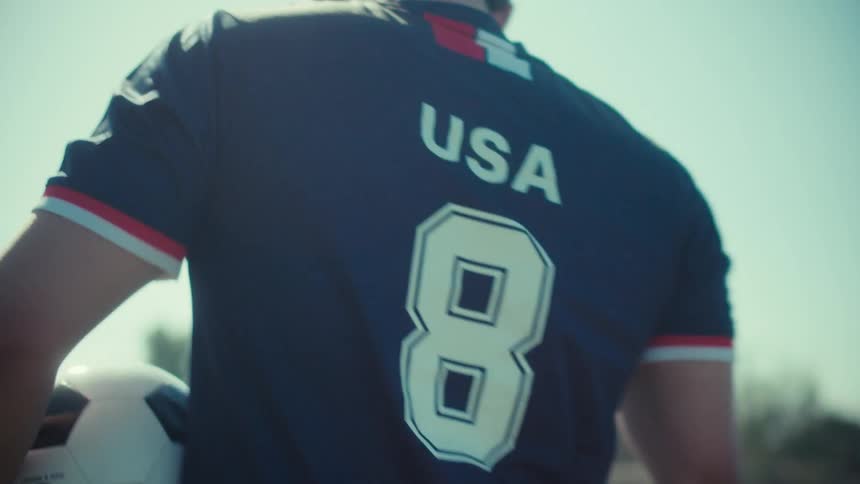 In slow motion, a determined male soccer player in a USA jersey walks onto the field with a soccer ball as the sun flares behind him
