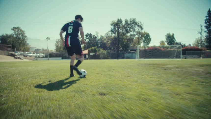 Dynamic follow shot of a male soccer player dribbling a ball across a sunny field in slow motion