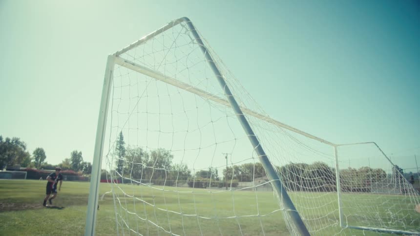 From behind the goal, a soccer ball soars into the net in slow motion on a bright day