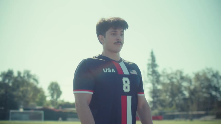 A focused young soccer player with a mustache stands proudly in a USA jersey on a sunny field, taking a deep breath