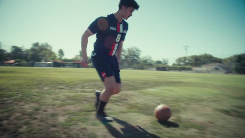 A young male athlete in a USA soccer jersey dribbles and kicks a vintage ball across a field in dynamic slow motion