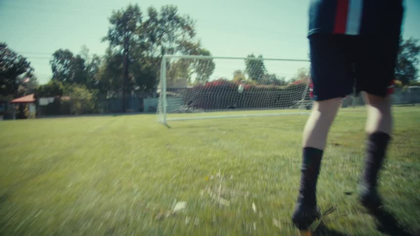 Dynamic low-angle follow shot of a soccer player dribbling the ball down a sunny field and scoring a goal