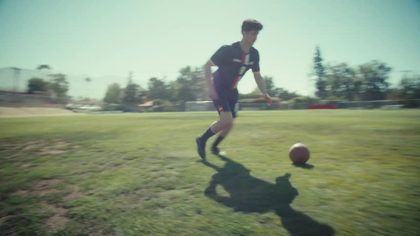 Slow motion tracking shot of a young male soccer player dribbling a ball on a sunny green field
