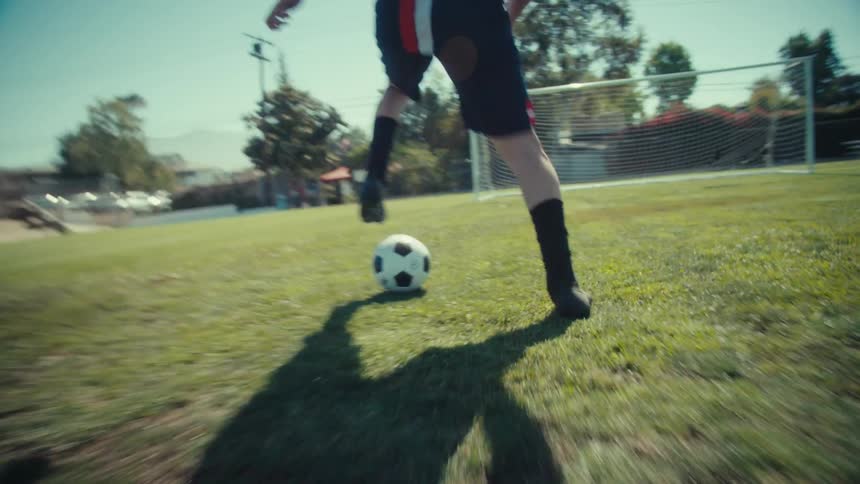 Dynamic low-angle tracking shot follows a soccer player's feet as he dribbles a ball down a sunny field in slow motion