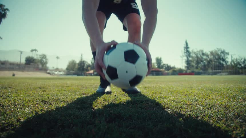 A soccer player in uniform walks across a sunny field in slow motion and places a ball on the grass to prepare for a penalty kick