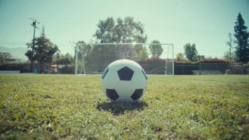A soccer player's foot kicks a ball on a green field towards an empty goal on a sunny day