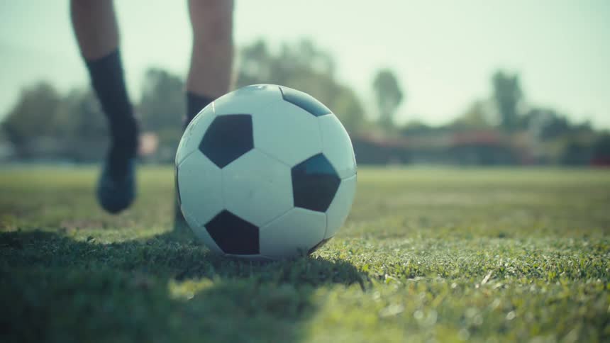 From a low angle on a sunny day, a soccer player carefully places a ball on the grass field, backs up, and takes a powerful kick