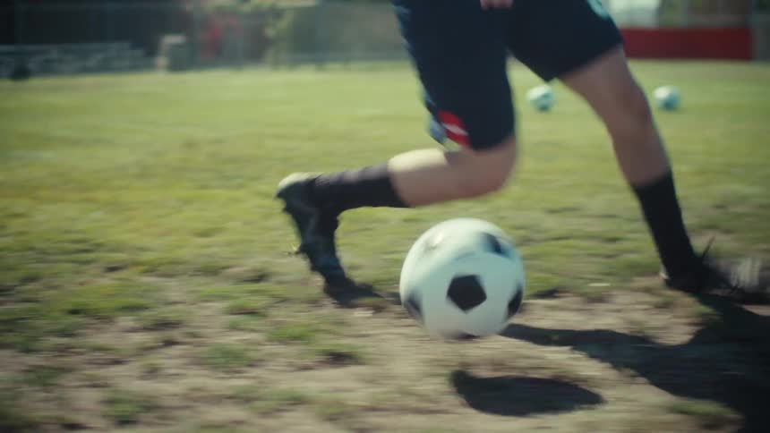 A soccer player skillfully dribbles a ball across a sunny field in a dynamic, low-angle slow-motion tracking shot