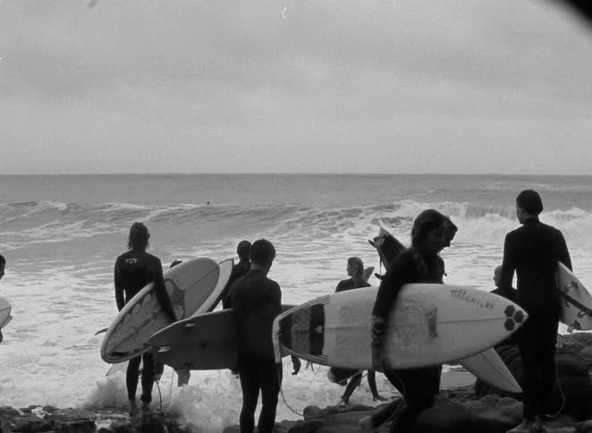 Vintage black and white footage shows a group of surfers with their boards contemplating the ocean waves from a rocky shore