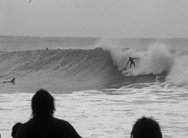 A grainy black and white shot shows a surfer skillfully riding a large, powerful ocean wave as spectators watch from the shore
