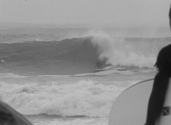 A grainy black and white slow-motion shot shows a surfer watching from shore as another surfer rides a large, powerful ocean wave