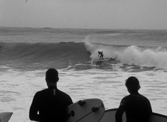 Grainy black and white footage shows silhouetted surfers watching another person ride a wave
