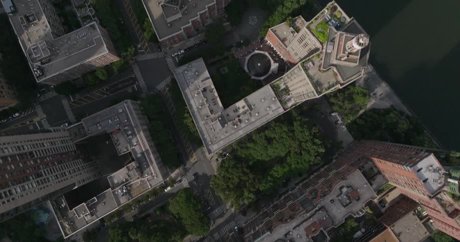 A top-down aerial view slowly rises over a dense urban neighborhood, showcasing the architecture of rooftops and green spaces alongside a river