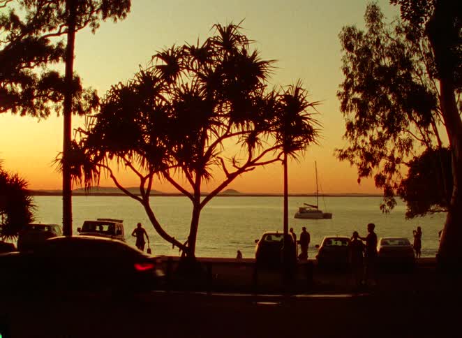 A tranquil tropical coastline at sunset with silhouettes of people, trees, and cars by the water