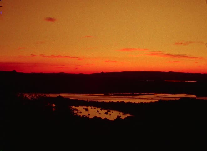 A vibrant, fiery sunset casts a deep orange and red glow across the sky, reflecting in the water below a silhouetted mountain range