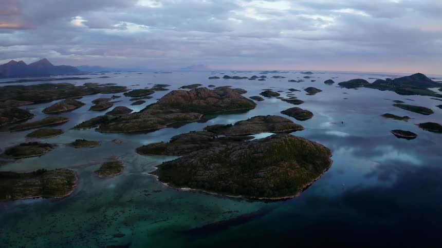 A static aerial view captures a vast archipelago of rocky islands in calm, turquoise waters under a dramatic, cloudy sky at dusk