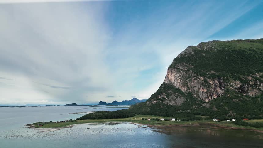 A majestic aerial view of a rugged green mountain overlooking a calm fjord and a small coastal village under a wide, cloudy blue sky