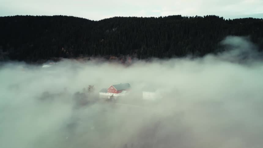 An aerial shot slowly pushes in on a red house surrounded by a sea of fog at the base of a forested mountain