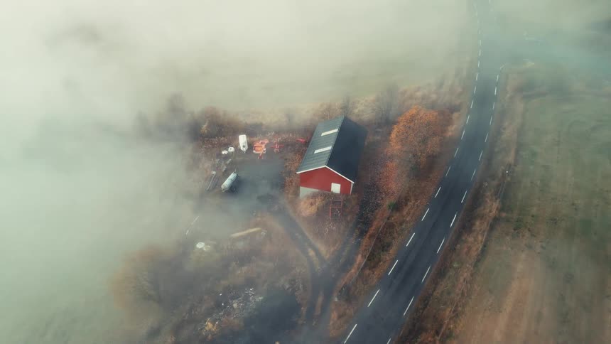 An aerial view shows a lone red barn next to a rural road as thick fog rolls over the landscape