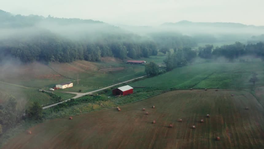 An early morning aerial shot flies over a field of hay bales towards a red barn nestled in a beautiful, foggy mountain valley