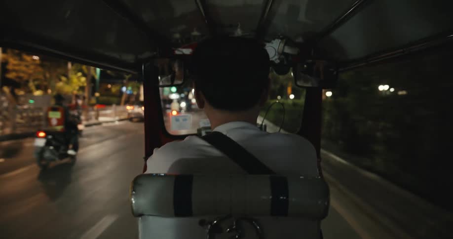 A passenger's point of view from the back of a tuk-tuk as it drives through the city streets at night