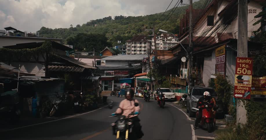 A winding road in a bustling Thai town sees a steady flow of motorcycle traffic on a cloudy day