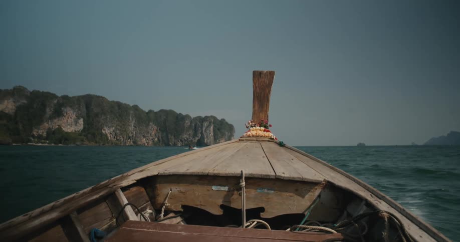 A point-of-view from the bow of a traditional wooden longtail boat as it travels across the ocean towards a tropical island with limestone cliffs