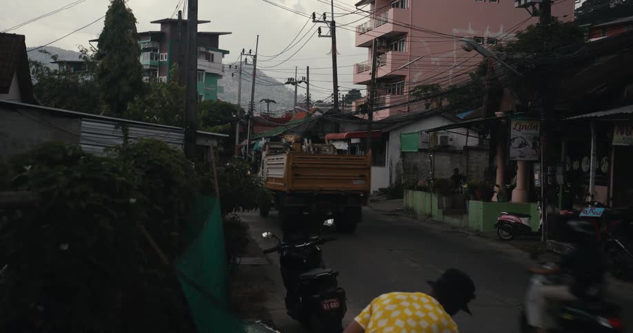 A yellow truck drives down a narrow city street in Thailand past apartment buildings and storefronts