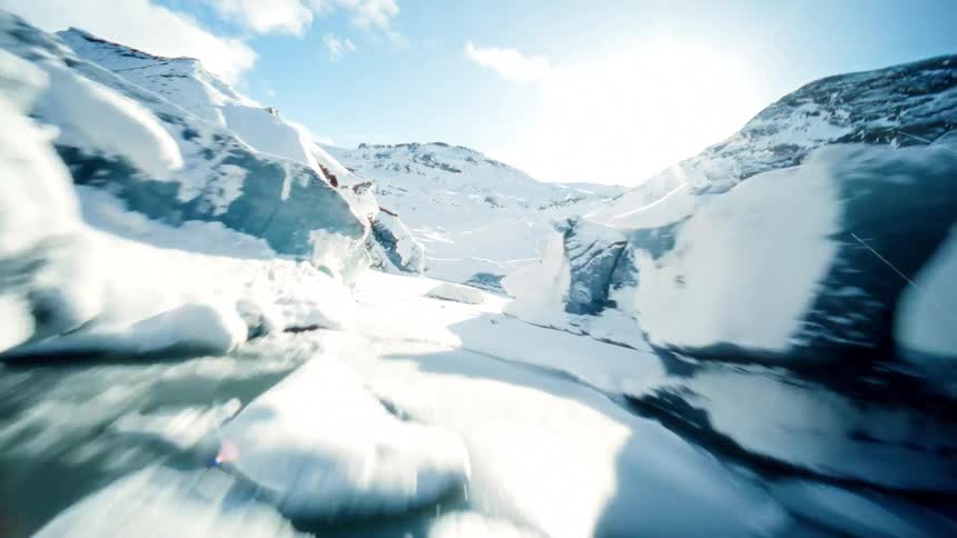 A thrilling FPV drone flight speeds over a vast glacier, swooping through a narrow blue ice cave under a bright sun