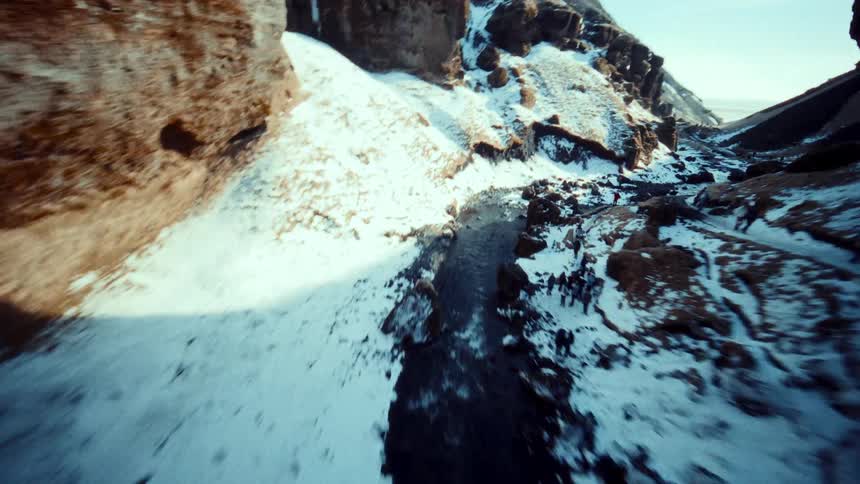 An exhilarating FPV drone flight dives over a majestic waterfall into a deep, snow-covered canyon on a bright winter day