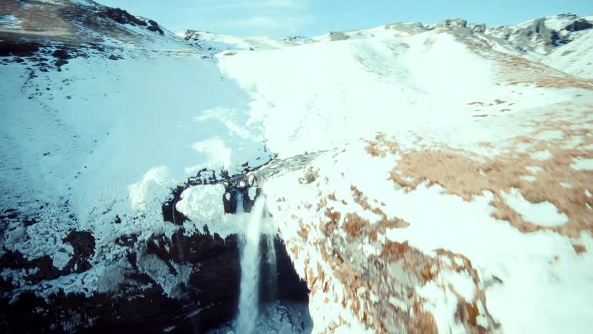 Dynamic FPV drone footage swoops down a dramatic, snow-covered canyon, flying over a river to a stunning Icelandic waterfall in winter