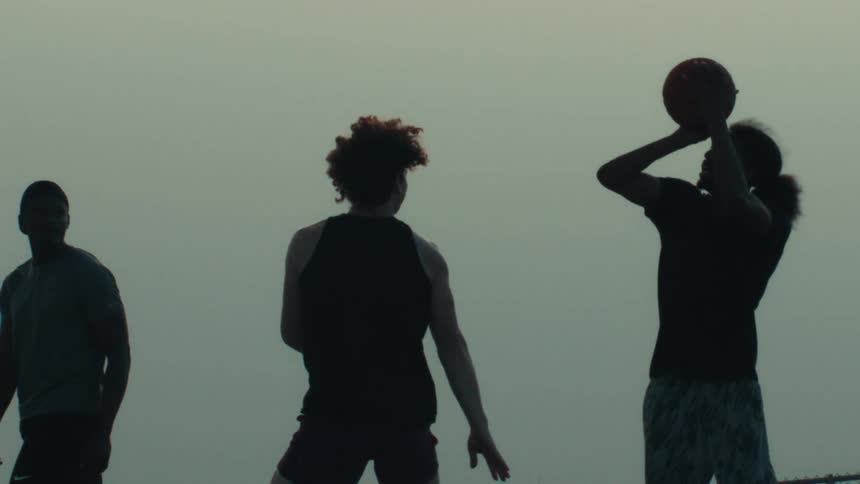 A silhouetted basketball player takes a dramatic slow-motion shot against a defender at dusk