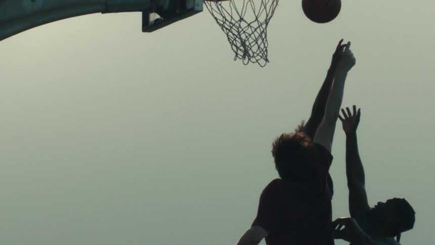 A slow-motion silhouette of basketball players jumping up to dunk the ball against a hazy sky