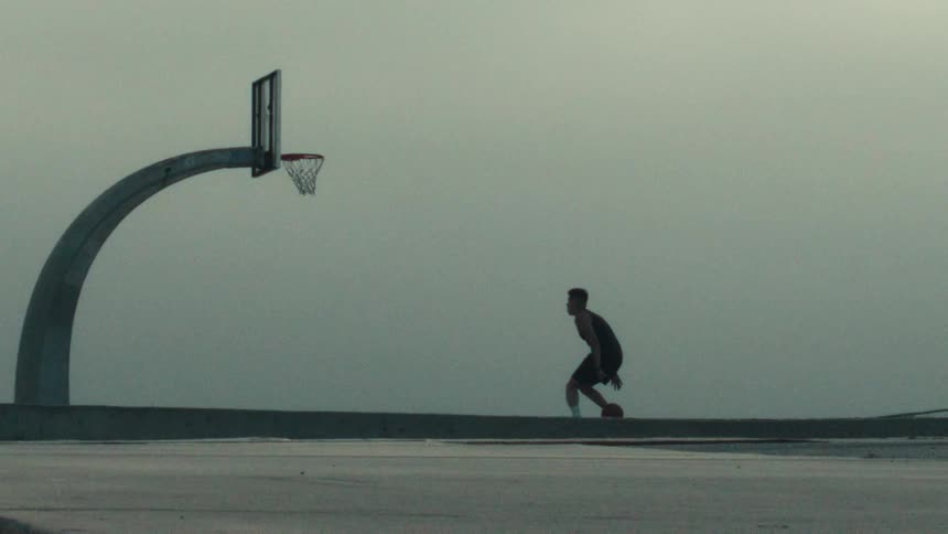 A silhouetted basketball player practices dribbling in slow motion on an outdoor court