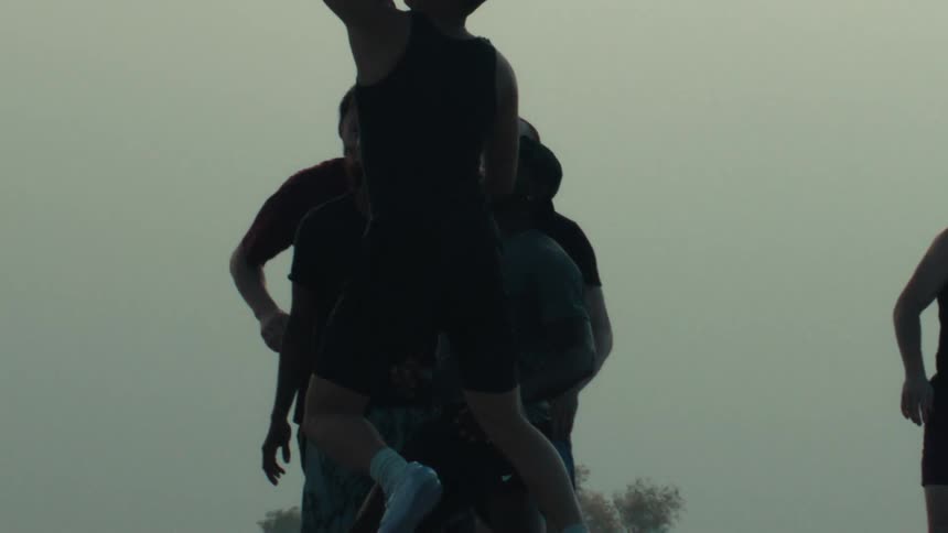 In dramatic slow motion, a silhouetted basketball player takes a shot as his teammates watch against a hazy sky