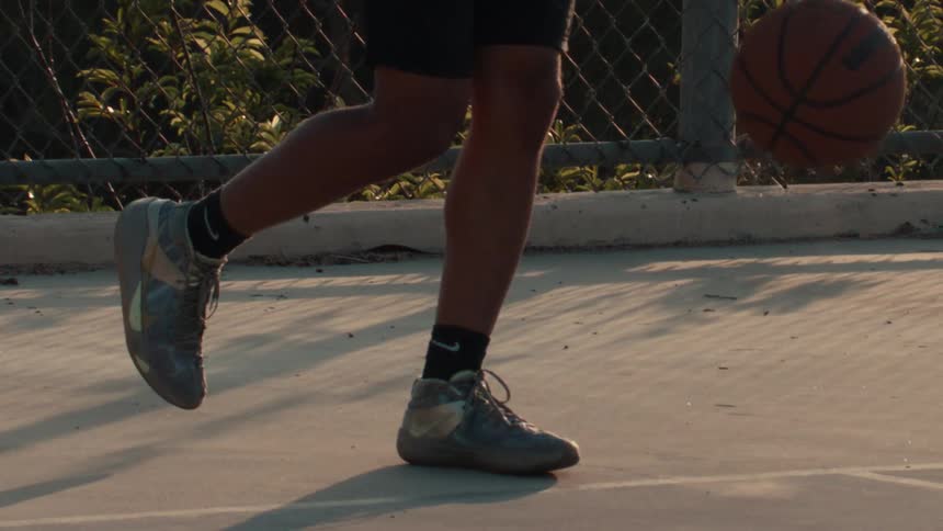 Close-up slow-motion footage captures a basketball player dribbling on an outdoor court at golden hour