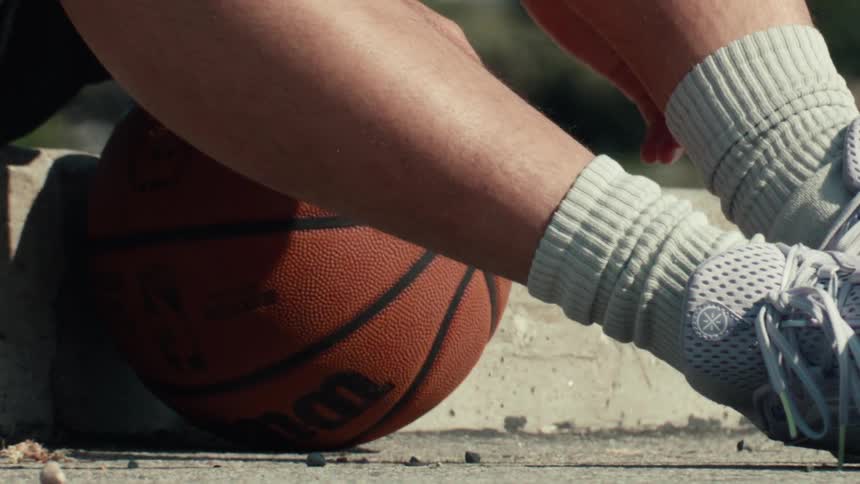 Slow motion close-up of a basketball player stretching their leg next to their basketball on a sunny day