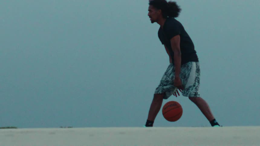 A focused man with curly hair dribbles a basketball in extreme slow motion on a sandy court at dusk