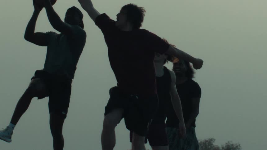 A group of young men play a dramatic game of basketball, silhouetted against the sky in slow motion