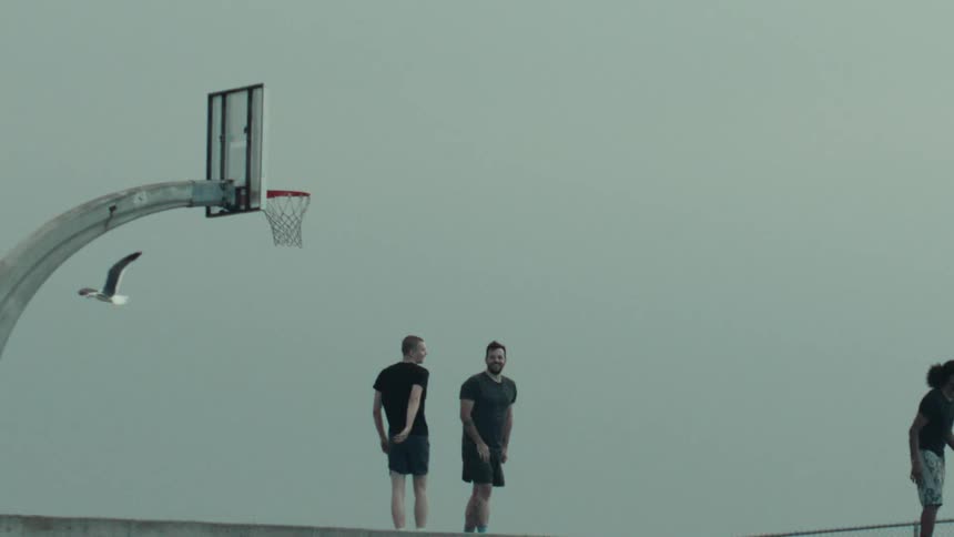 A cinematic slow motion view of a seagull flying past three men standing on an outdoor basketball court under a gray sky