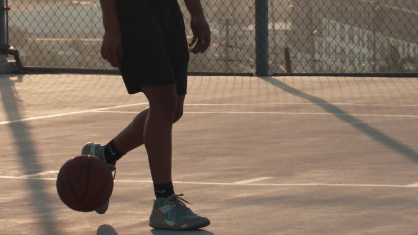 Low angle slow-motion shot of a basketball player's feet dribbling a ball on an outdoor court at sunset