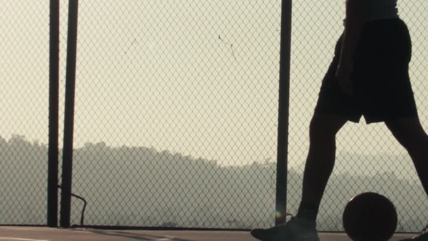 Low angle slow motion shot of a basketball player's silhouette dribbling a ball on an outdoor court at golden hour