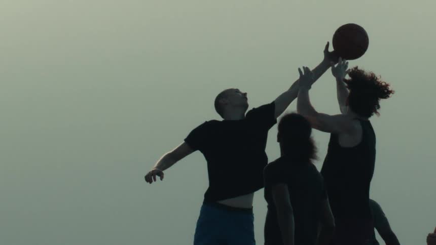 In a dramatic slow-motion silhouette, a group of young men leap to catch a basketball against the hazy sky