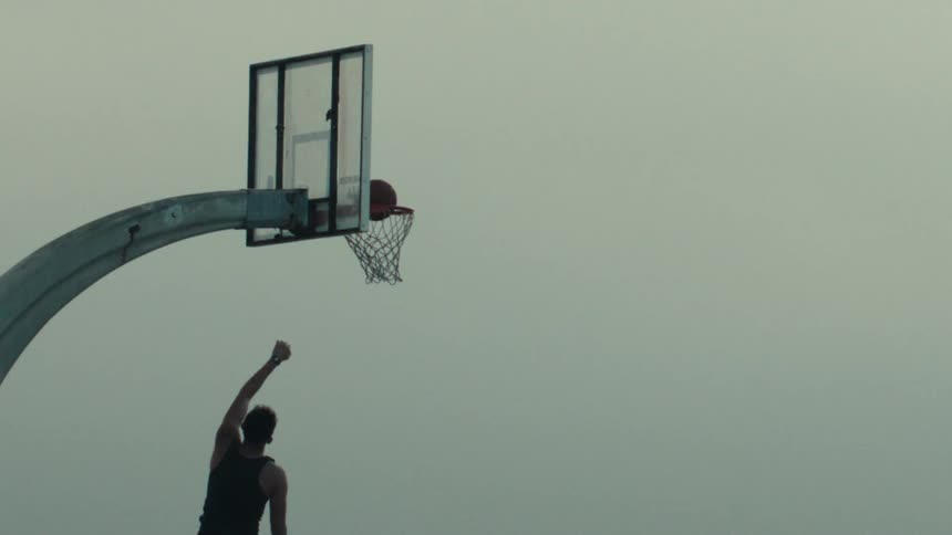 An athletic man dunks a basketball in dramatic slow motion against an overcast sky