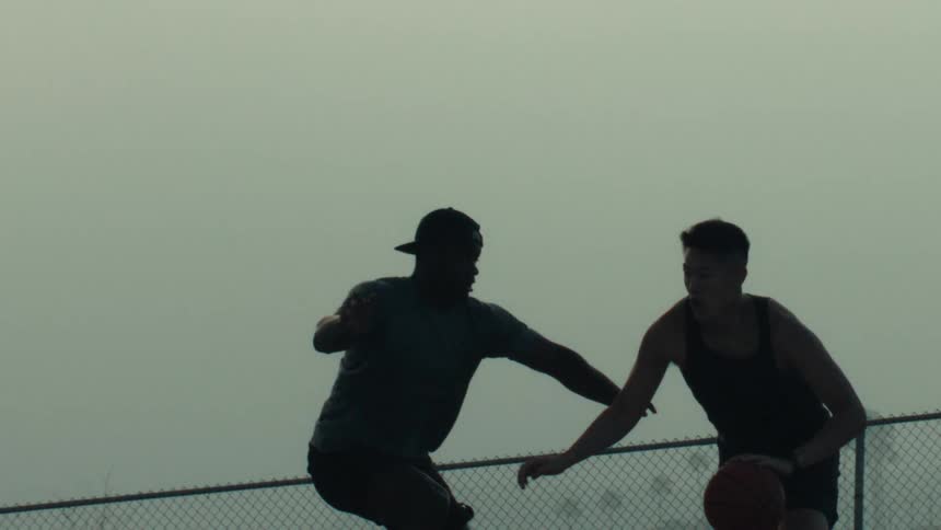 Friends compete in a game of one-on-one basketball, silhouetted against the sky in dramatic slow motion