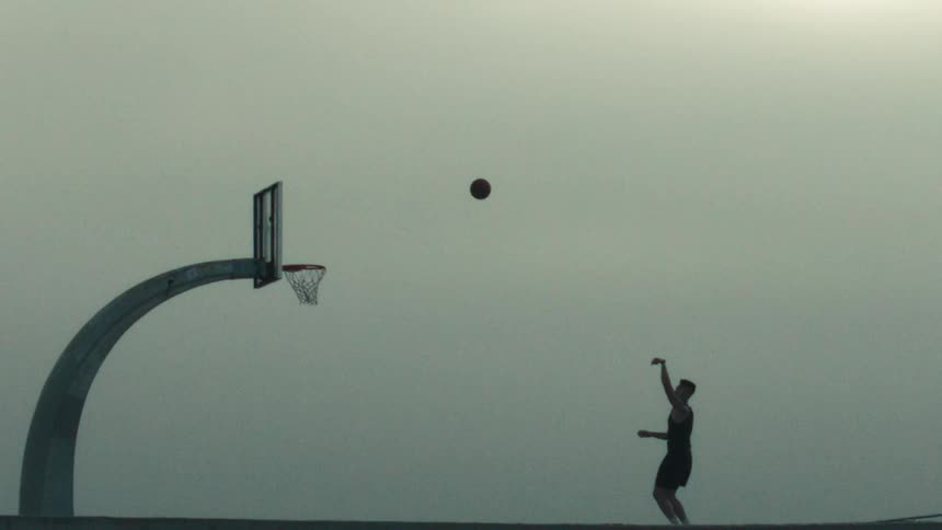 A basketball player's silhouette is captured in slow motion as they make a perfect shot into the hoop against a foggy sky