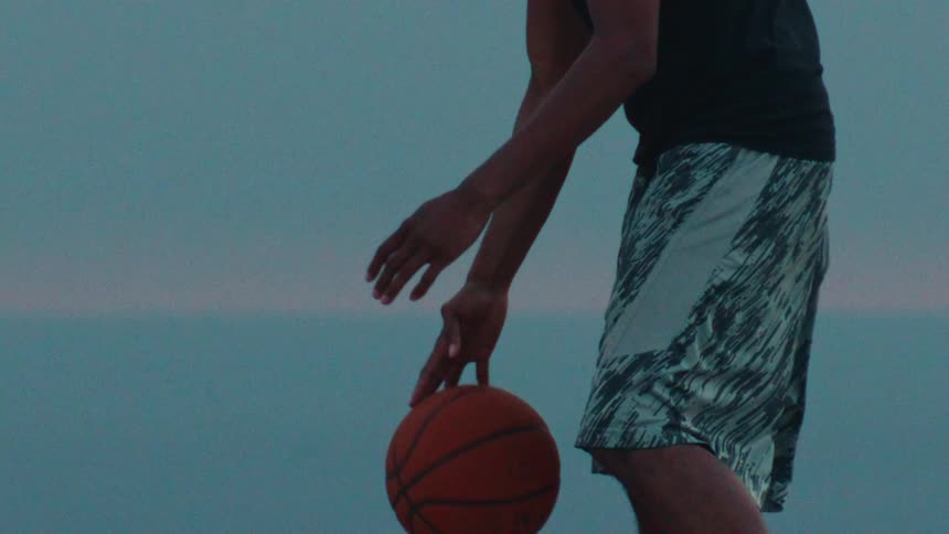 A basketball player practices dribbling moves in super slow motion against a serene ocean backdrop at dusk
