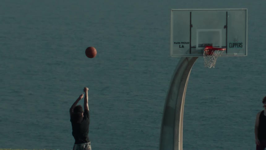 A person shoots a basketball in slow motion on an outdoor court with a stunning ocean view in the background